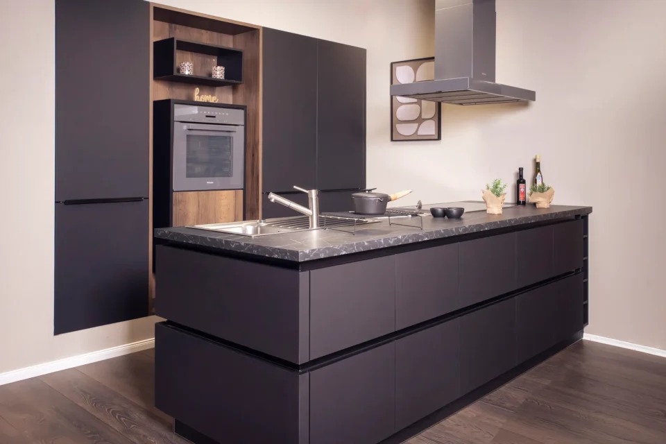 Moderne Küche mit dunklen Schränken, zentraler Insel mit Spüle und Herd, und eingebautem Backofen. Küchenutensilien und Dekoartikel, wie Pflanzen und Flaschen, sind auf der Arbeitsplatte platziert. Wandbild im Hintergrund.