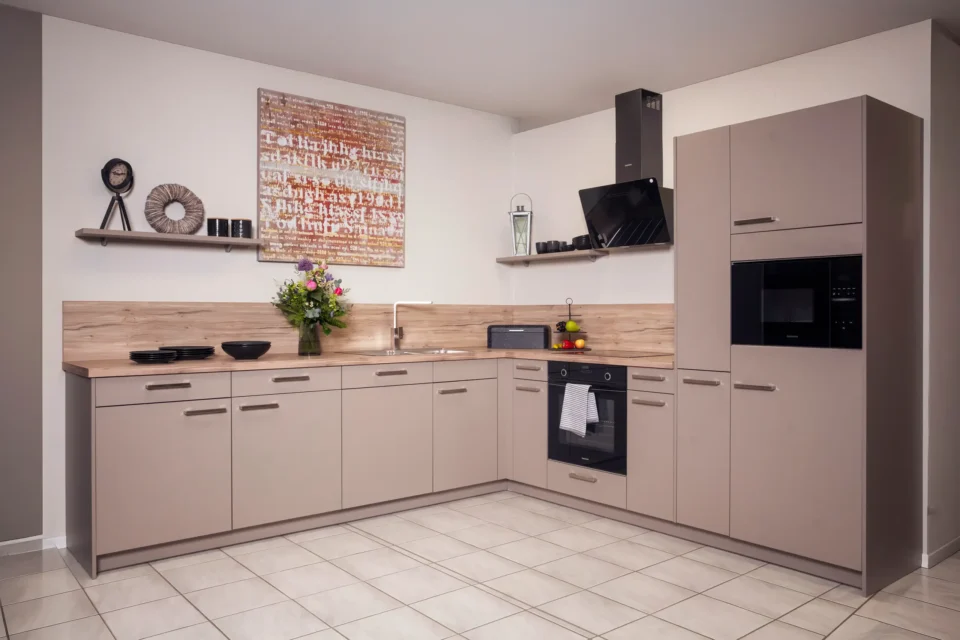 Moderne Küche mit L-förmigen, hellbraunen Schränken, hellem Holzarbeitsbereich und schwarzem Herd. An der Wand sind Regale mit Dekoration. Ein Strauß Blumen steht auf der Arbeitsfläche, Fliesenboden in hellem Beige.