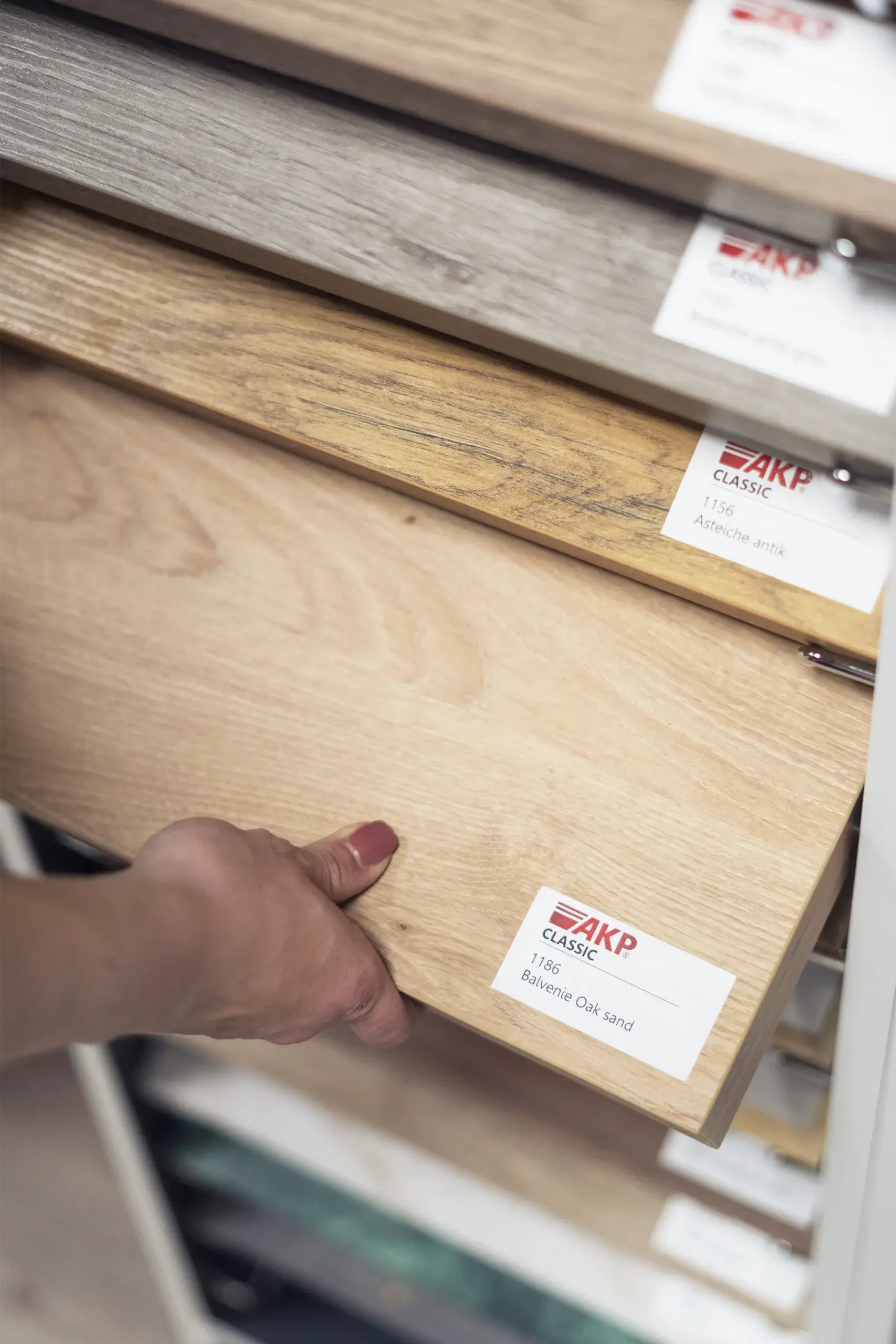 Kuechenstudio De Koeoek Kuechenausstellung Musterauswahl Eine Hand zieht ein Musterbrett aus Holzoptik aus einem Regal mit mehreren Holzmusterplatten. Jede Platte hat ein Etikett mit der Aufschrift "AKP Classic" und anderen Details.