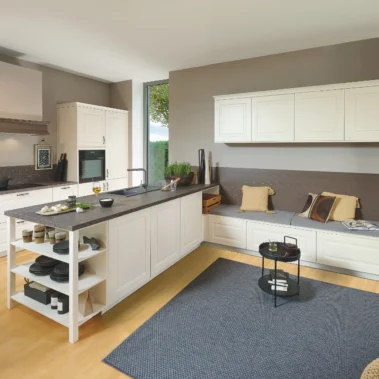 Eine moderne Küche mit weißem Mobiliar, Arbeitsflächen aus dunklem Stein und offenem Regal. Links ein Ofen, rechts ein Sitzbereich mit Kissen und eingebauten Regalen. Heller Holzboden, grauer Teppich und große Fenster mit Blick ins Freie.