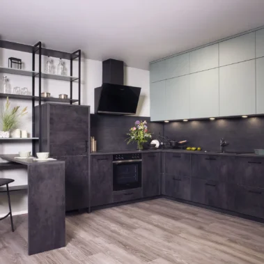Moderne Küche mit grauen Schränken und Geräten. Offene Regale mit Geschirr links, ein Stuhl am schmalen Tisch. Beleuchtung unter den Oberschränken, Blumen und Obstschalen auf der Arbeitsplatte. Holzboden.
