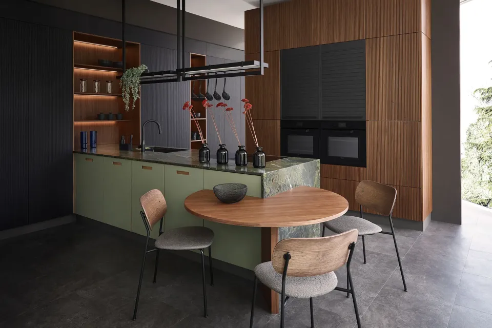 Moderne Küche mit Holz- und dunklen Akzenten. Eine Kücheninsel mit grünem Marmorsockel steht zentral, daneben ein runder Holztisch mit Stühlen. Einbauschränke und große Fenster sorgen für viel Licht.
