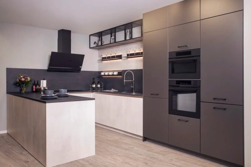 Moderne Küche mit grauen und weißen Schränken, dunkler Arbeitsplatte, eingebauten Geräten und Herd. Offene Regale mit Gläsern sind beleuchtet. Ein Blumenstrauß und Geschirr befinden sich auf der Kochinsel. Heller Holzboden.