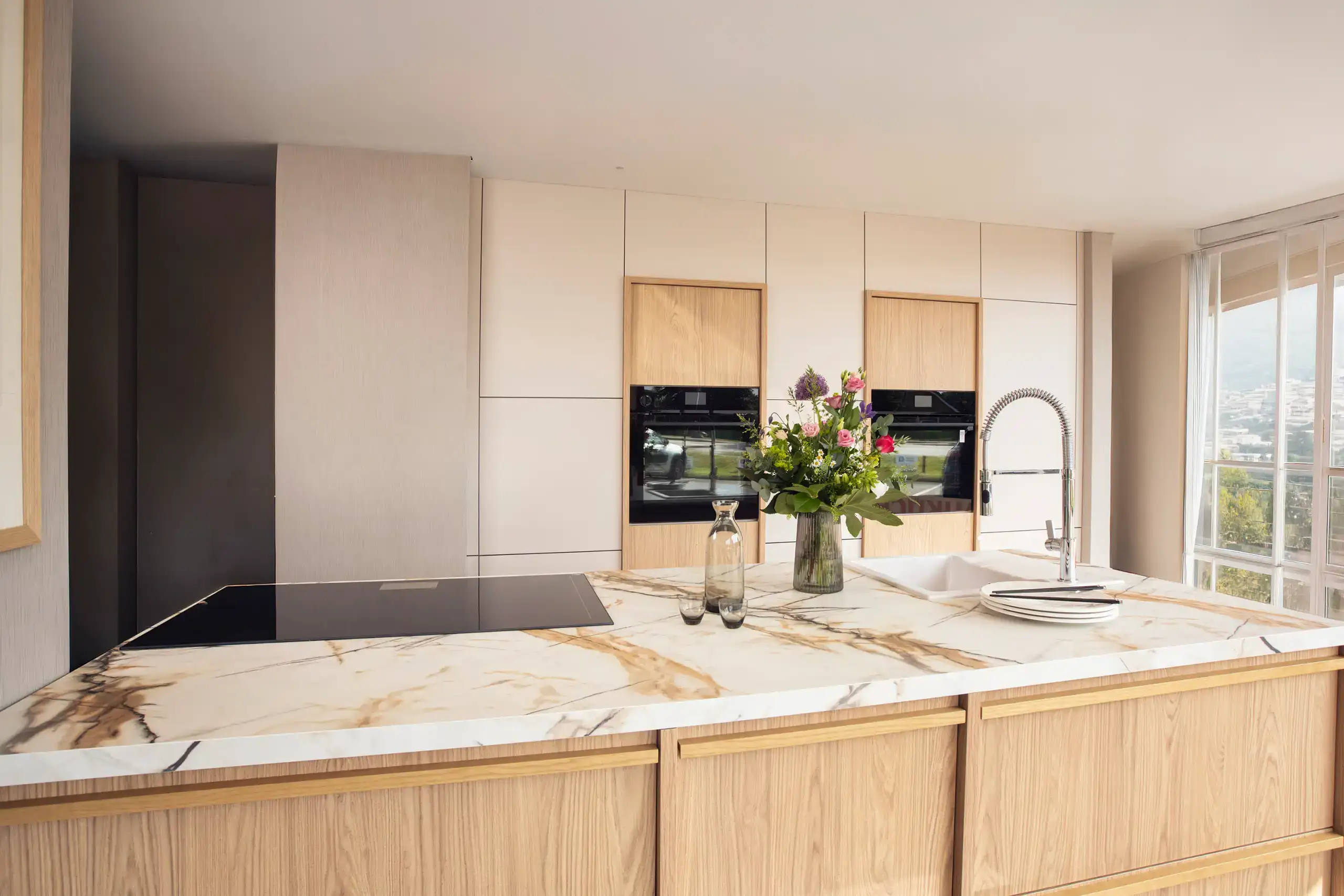 Moderne Küche mit hellen Holz- und Marmorelementen. Ein großer Küchenblock mit Induktionskochfeld und Spüle steht im Vordergrund. Darauf stehen Blumen in einer Vase und Geschirr. Im Hintergrund sind Einbauschränke mit integrierten Geräten.