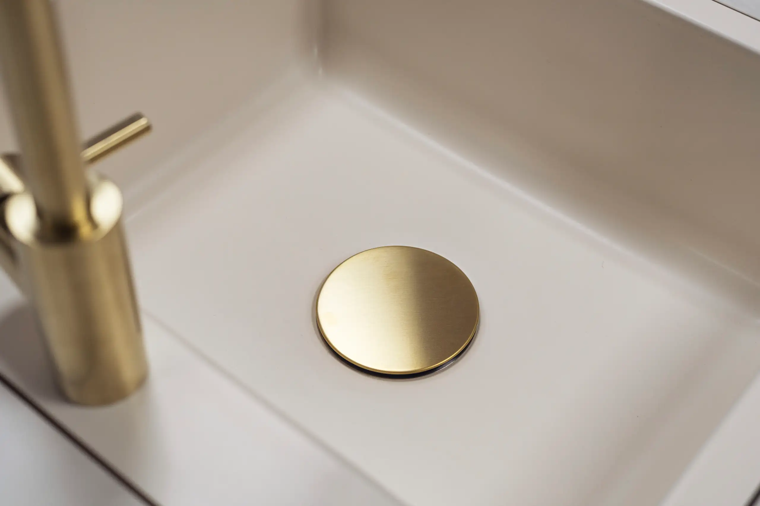 Ein Waschbecken mit einem runden, goldfarbenen Abflussdeckel. Der Wasserhahn und die Ränder des Waschbeckens sind ebenfalls sichtbar. Das Design wirkt modern und minimalistisch.