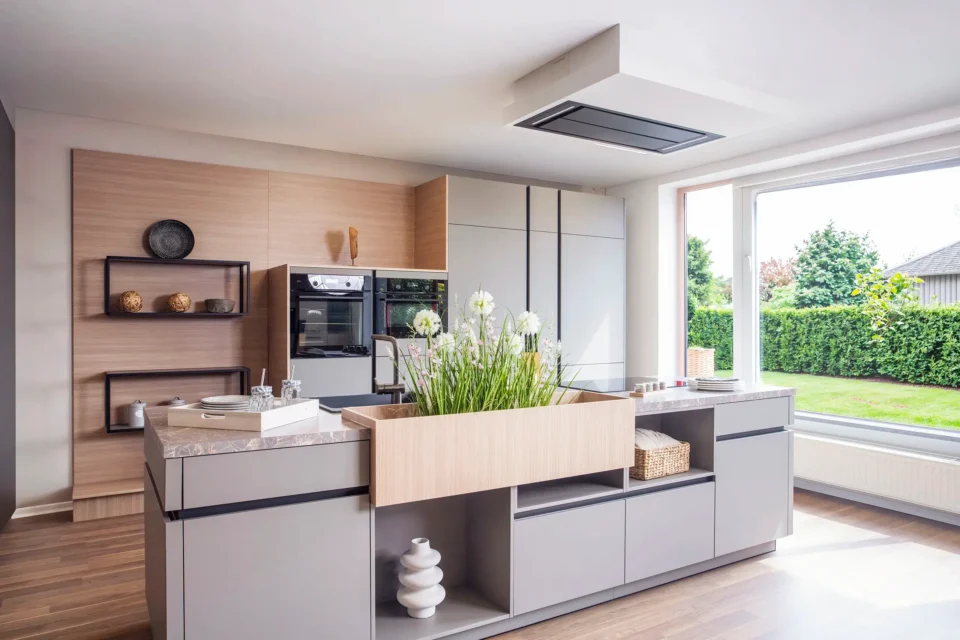 Moderne Küche mit grauen Schränken, zentralem Küchenblock mit integrierter Spüle und Pflanzen. Große Fenster lassen Tageslicht herein, Holzböden und Regale mit Dekorationselementen wie Körben und Vasen im Hintergrund.