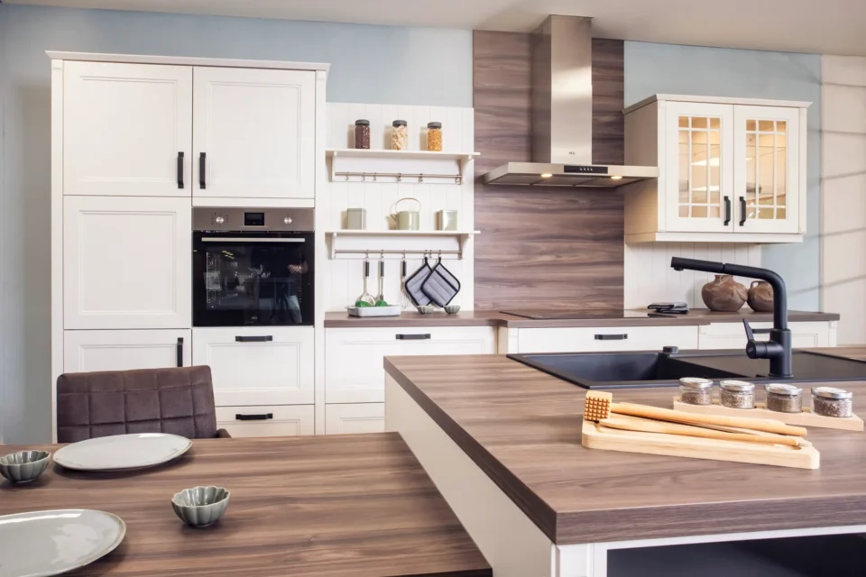 Moderne Küche mit weißem Schrank, eingebautem Ofen, Herd mit Dunstabzugshaube und brauner Arbeitsplatte. Auf der Theke stehen Gewürzdosen und Küchenutensilien. Ein Esstisch mit Tellern und Schalen ist im Vordergrund.