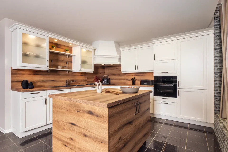 Moderne Küche mit weißen Schränken, Holzarbeitsplatten und einer zentralen Kücheninsel aus Holz. Die Rückwand ist im Holzdesign gestaltet. Schwarzer Backofen und Dekorationsgegenstände wie Pflanzen und Vasen ergänzen das Bild.