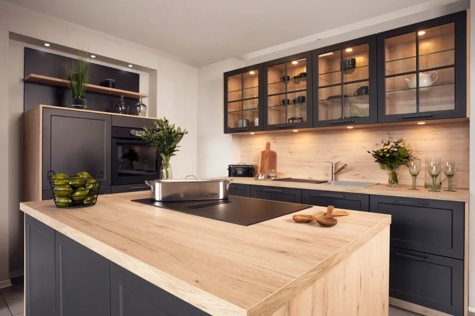 Eine moderne Küche mit einer zentralen Insel aus Holz und grau-schwarzen Akzenten. Ein eingebauter Ofen, dekorative Pflanzen, Gläser und Küchenutensilien sind sichtbar. Schränke haben beleuchtete Glasfronten.