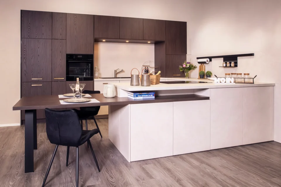 Eine moderne Küche mit dunklen Holzmöbeln und weißen Fronten. Esstisch angeschlossen, schwarze Stühle, Dekorationen und Bücher. Blumenstrauß auf der Arbeitsplatte, Regale an der Wand. Holzfußboden.