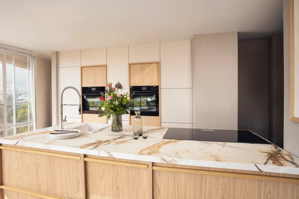 Moderne Küche mit hellen Holz- und weißen Schränken. In der Mitte befindet sich eine Insel mit Marmorarbeitsplatte, Spüle, Wasserhahn und Kochfeld. Darauf stehen ein Blumenstrauß und eine Flasche. Fenster im Hintergrund.