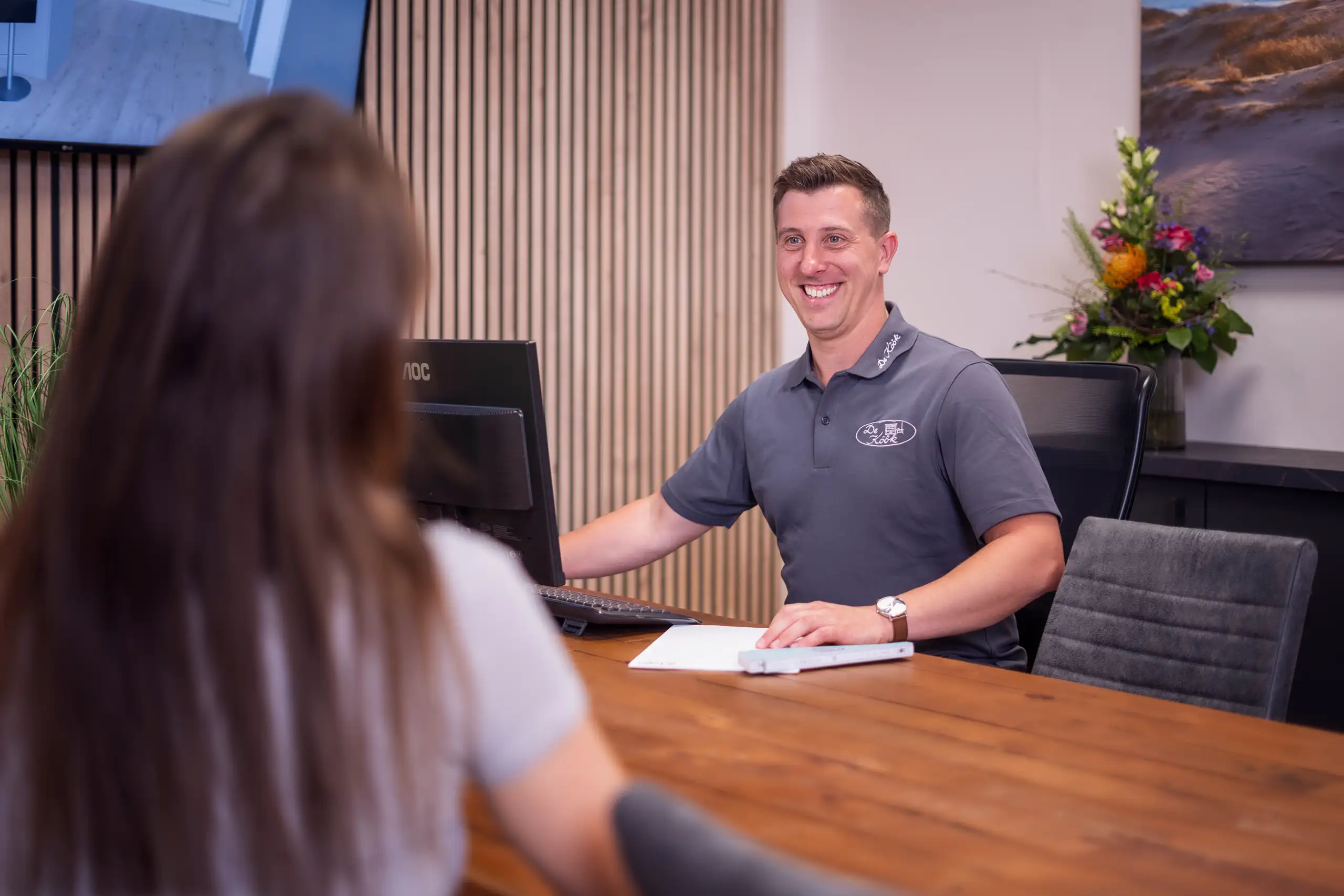 Ein Mann sitzt lächelnd an einem Holztisch mit einem Computerbildschirm und Unterhaltungstechnik. Er trägt ein graues Poloshirt. Im Hintergrund sind vertikale Holzlatten und ein Blumenarrangement zu sehen. Eine Person sitzt ihm gegenüber.