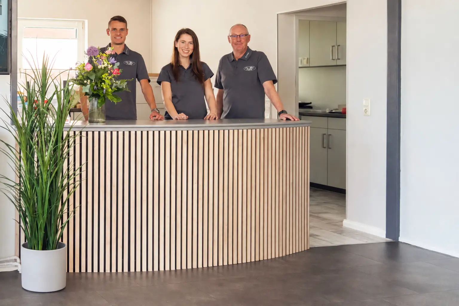 Drei Personen in einheitlichen grauen Poloshirts stehen hinter einem halbkreisförmigen Tresen mit Holzlamellen. Links steht eine Vase mit bunten Blumen, rechts eine Topfpflanze. Im Hintergrund ist ein Raum mit Küchenschränken erkennbar.