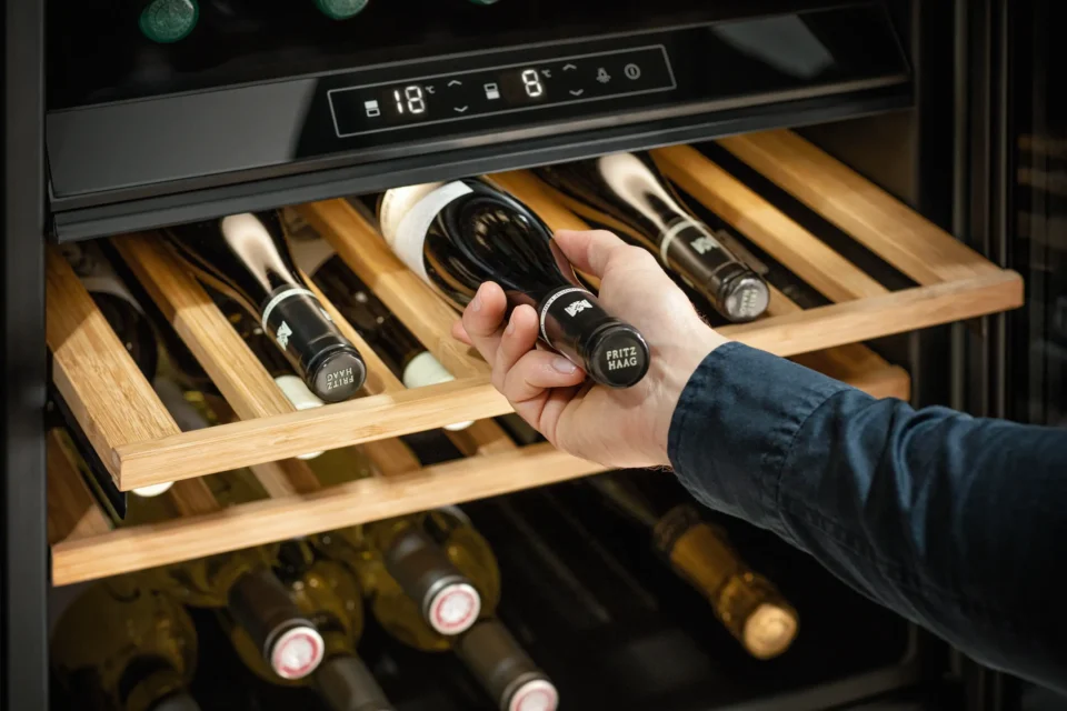 Eine Person zieht eine Weinflasche aus einem Kühlschrank mit Holzregalen. Die Temperatur des Kühlers beträgt 18 Grad. Weitere Weinflaschen sind in den Regalen darunter sichtbar.