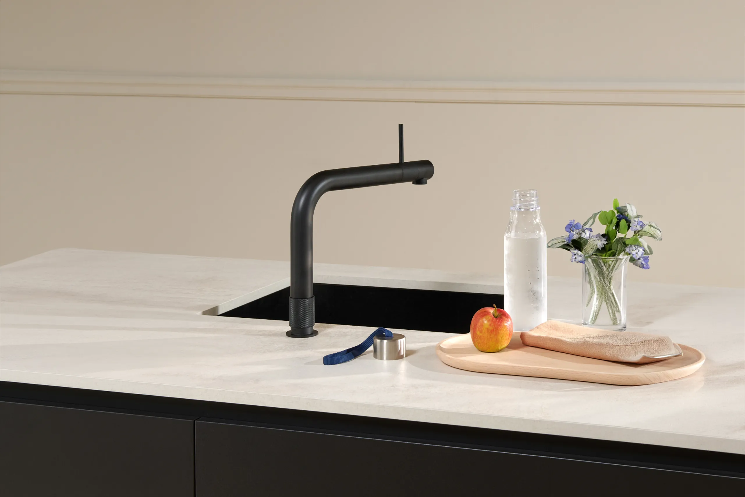 Eine moderne Küchenzeile mit einem schwarzen Wasserhahn vor einer beigen Wand. Auf der Arbeitsfläche liegen eine Trinkflasche, ein Apfel, ein blaues Armband, ein Schneidebrett sowie eine Vase mit Blumen.