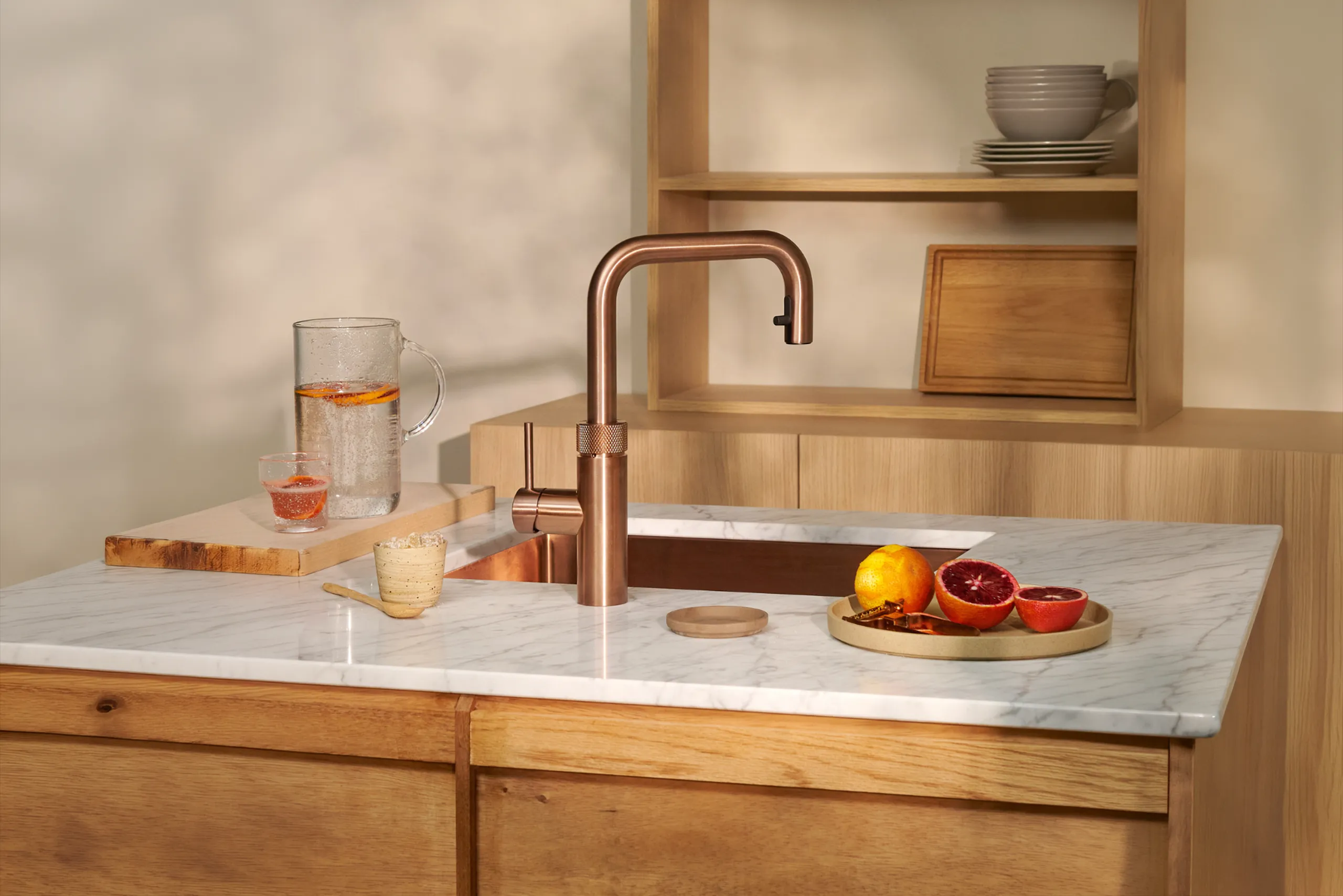 Eine Küche mit einer weißen Marmor-Arbeitsplatte. Darauf ein kupferfarbener Wasserhahn, ein Glas mit Orangenscheiben gefüllter Krug, ein Glas Wasser, eine Kerze und ein Teller mit Orangen und Gewürzen. Regal im Hintergrund.