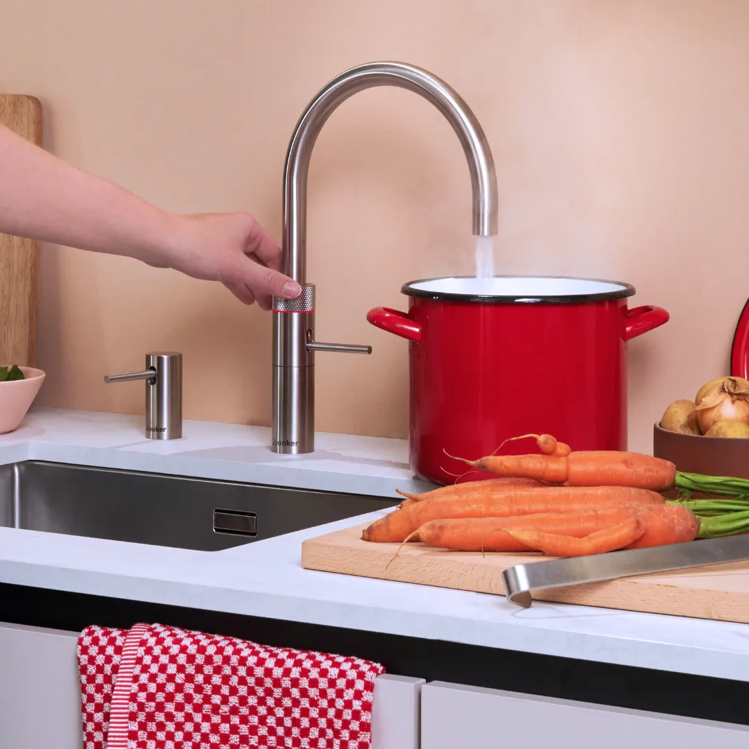 Eine Hand betätigt einen modernen Wasserhahn, aus dem Wasser in einen roten Topf läuft. Auf der Arbeitsplatte liegen Karotten und ein Geschirrtuch mit rotem Muster, daneben eine Schüssel mit Gemüse.