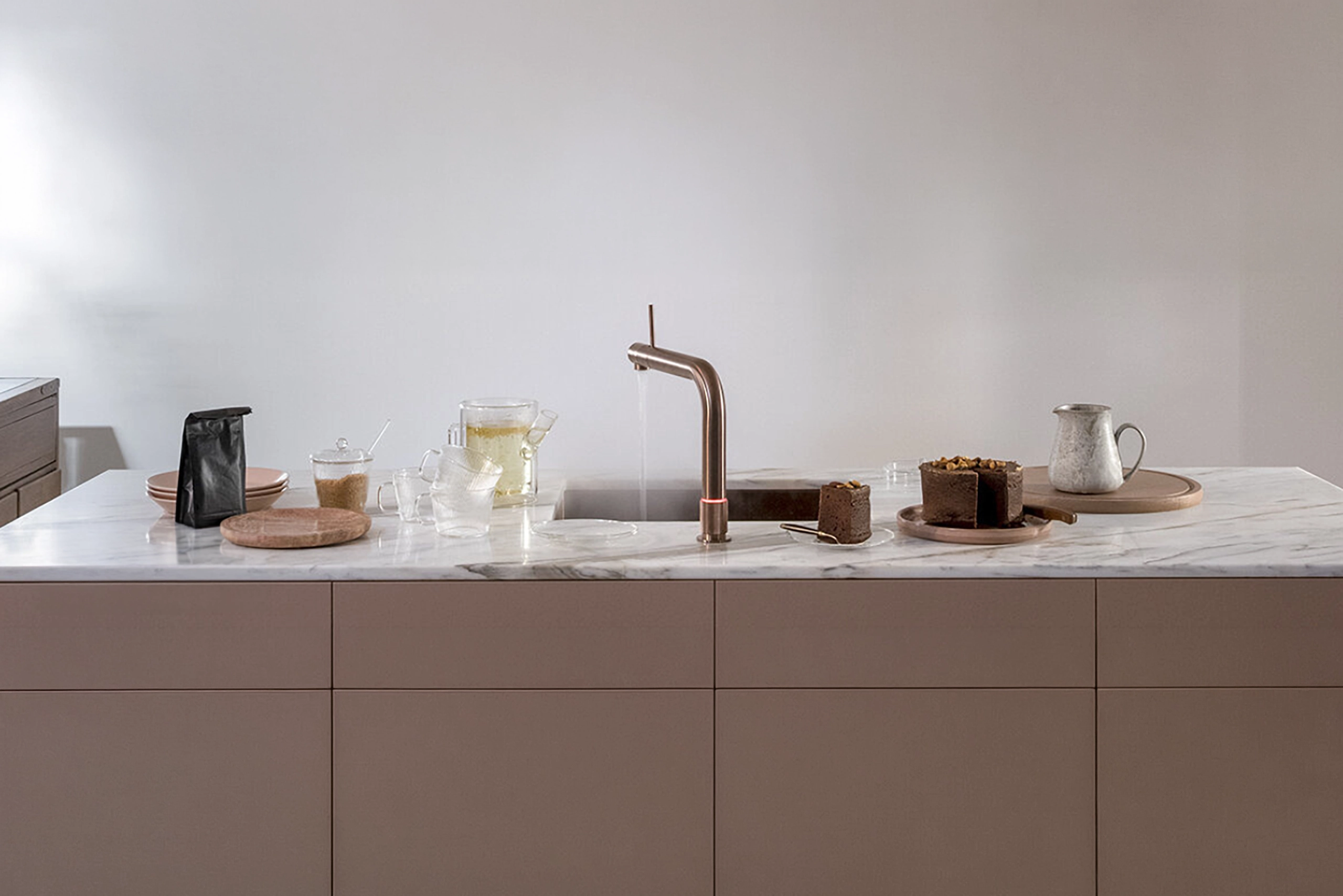 Eine minimalistische Küchenzeile mit einer Marmor-Arbeitsplatte. Darauf stehen eine schwarze Tüte, ein Schneidebrett, eine Stablampe, ein Wasserhahn, ein Glas Tee, ein Kuchen und ein Krug.