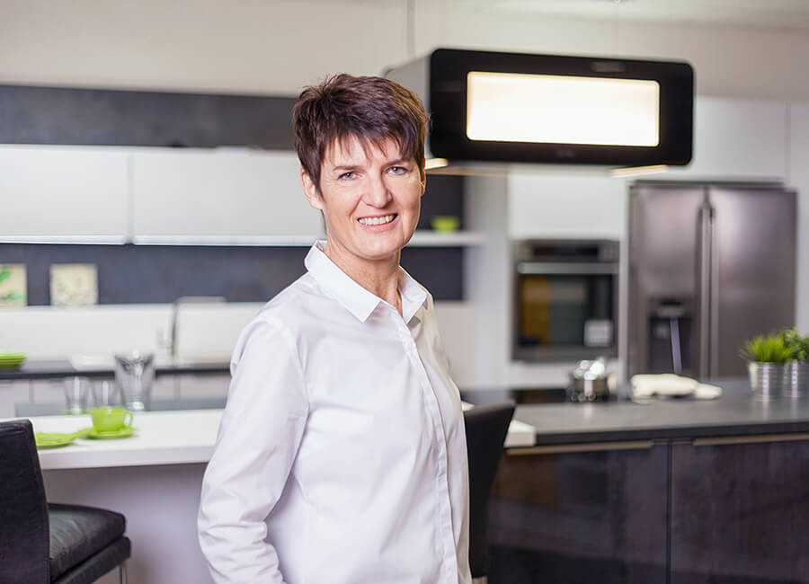 Eine Frau mit kurzen, dunklen Haaren steht in einer modernen Küche. Sie trägt ein weißes Hemd und lächelt in die Kamera. Im Hintergrund sind Küchenschränke, ein Ofen und ein Kühlschrank zu sehen.